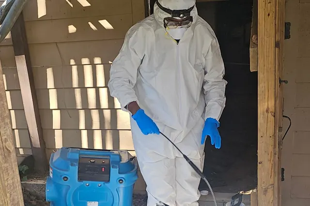 Fresh crew member in a protective suit spraying treatment beside a blue air mover
